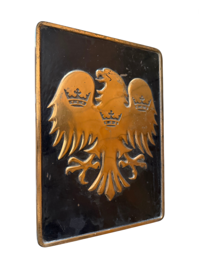 A rectangular black plaque featuring a raised gold double-headed eagle with three crowns, photographed against a plain white background.