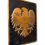 A rectangular black plaque featuring a raised gold double-headed eagle with three crowns, photographed against a plain white background.
