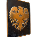 A rectangular black plaque featuring a raised gold double-headed eagle with three crowns, photographed against a plain white background.