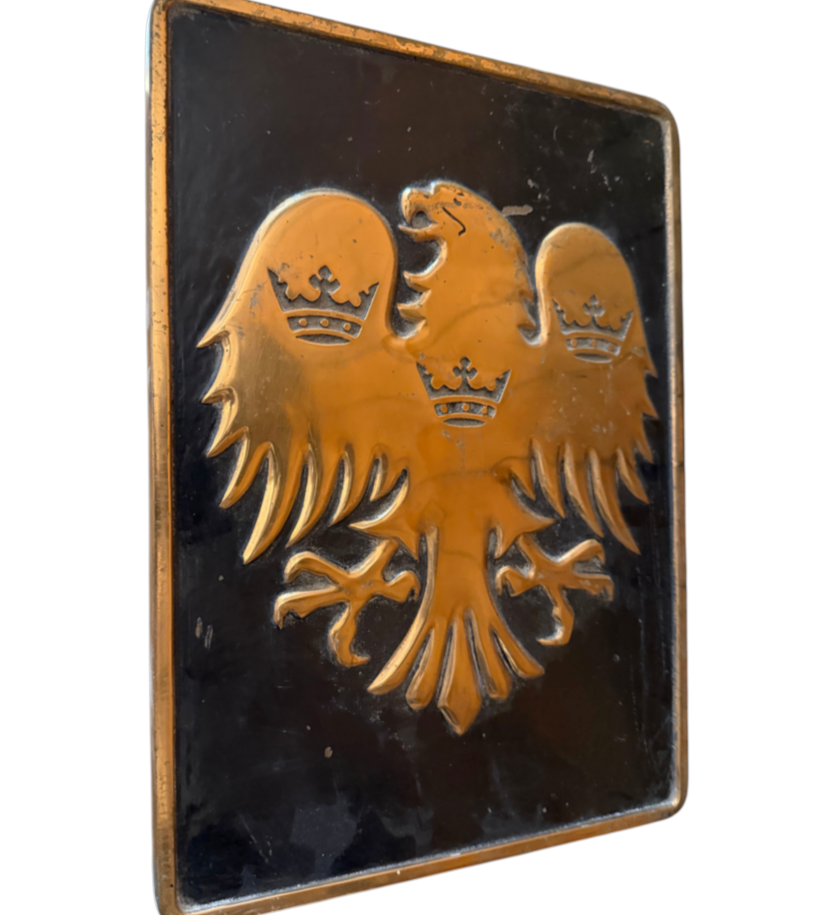 A rectangular black plaque featuring a raised gold double-headed eagle with three crowns, photographed against a plain white background.